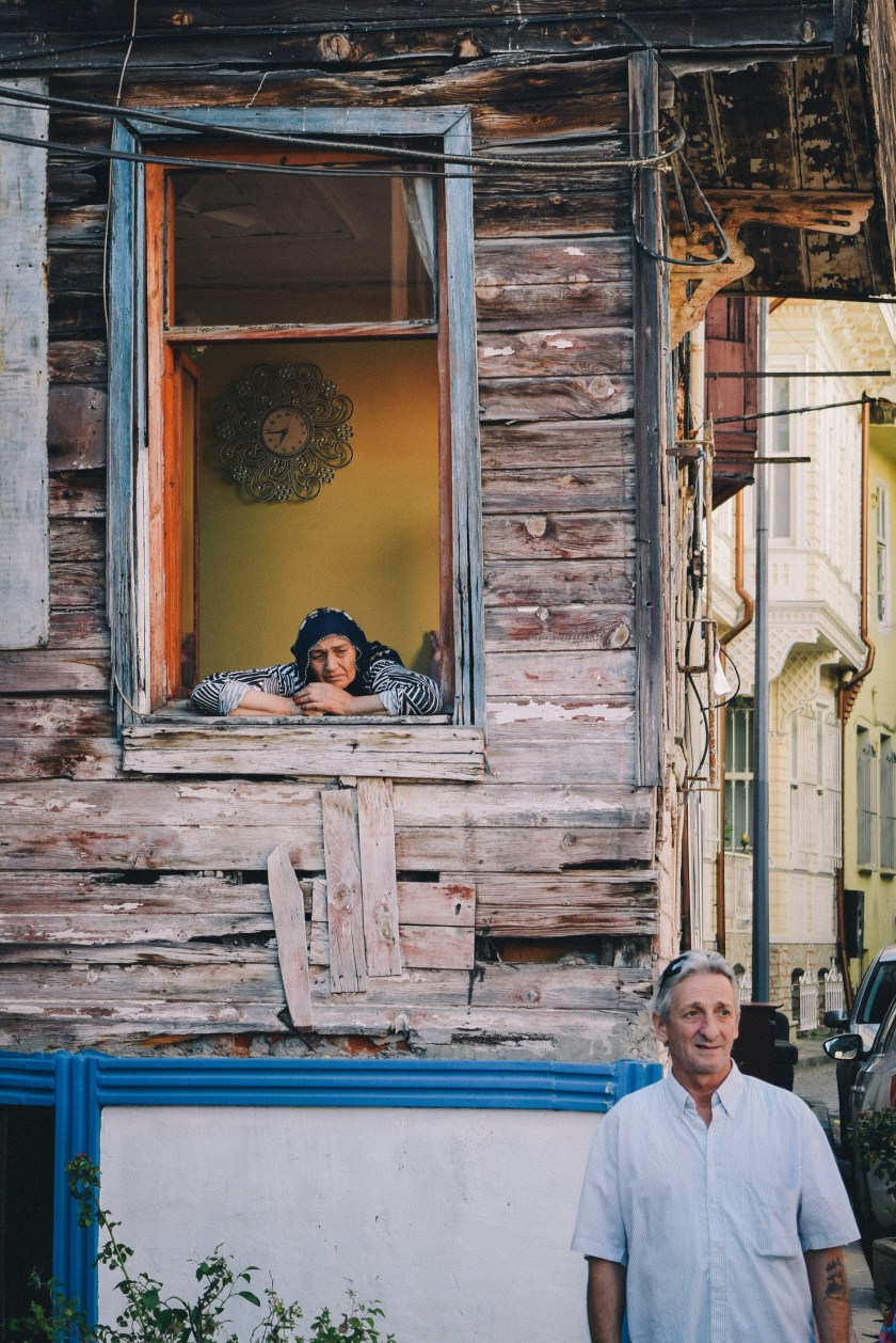 O femeie în vârstă stă rezemată în rama unui geam deschis al unei case vechi din lemn, cu pereți degradați. În interior, pe peretele galben, se vede un ceas ornamental. Sub fereastră, un bărbat mai în vârstă, îmbrăcat într-o cămașă deschisă la culoare, privește în lateral și zâmbește ușor. Scena surprinde un moment calm într-un cartier vechi.