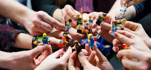 Several people’s hands gathered in a circle, each holding a small LEGO minifigure. The figures represent different characters and roles, symbolizing diversity, collaboration, and shared participation.