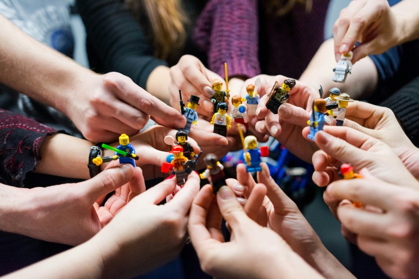 Several people’s hands gathered in a circle, each holding a small LEGO minifigure. The figures represent different characters and roles, symbolizing diversity, collaboration, and shared participation.